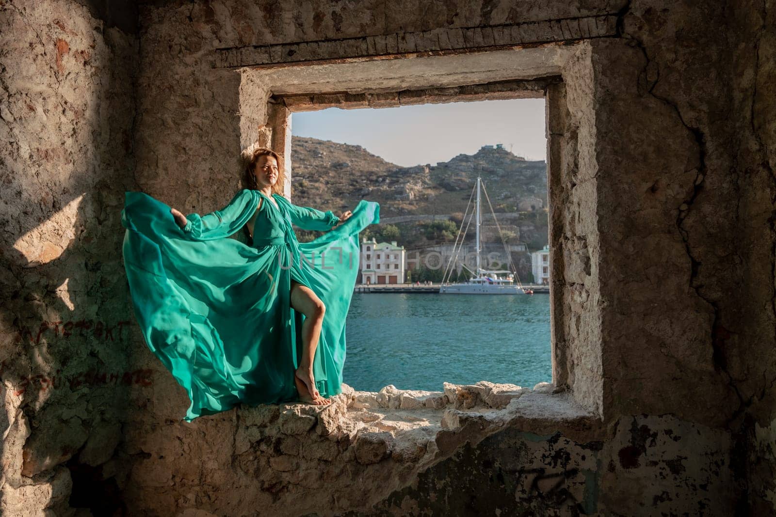 Rear view of a happy blonde woman in a long mint dress posing against the backdrop of the sea in an old building with columns. Girl in nature against the blue sky