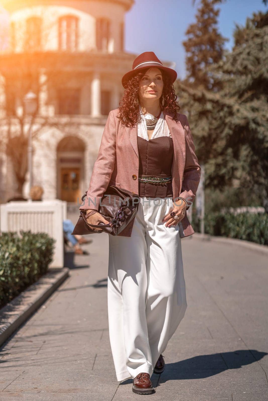 Woman park city. Stylish woman in a hat walks in a park in the city. Dressed in white corset trousers and a pink jacket with a bag in her hands