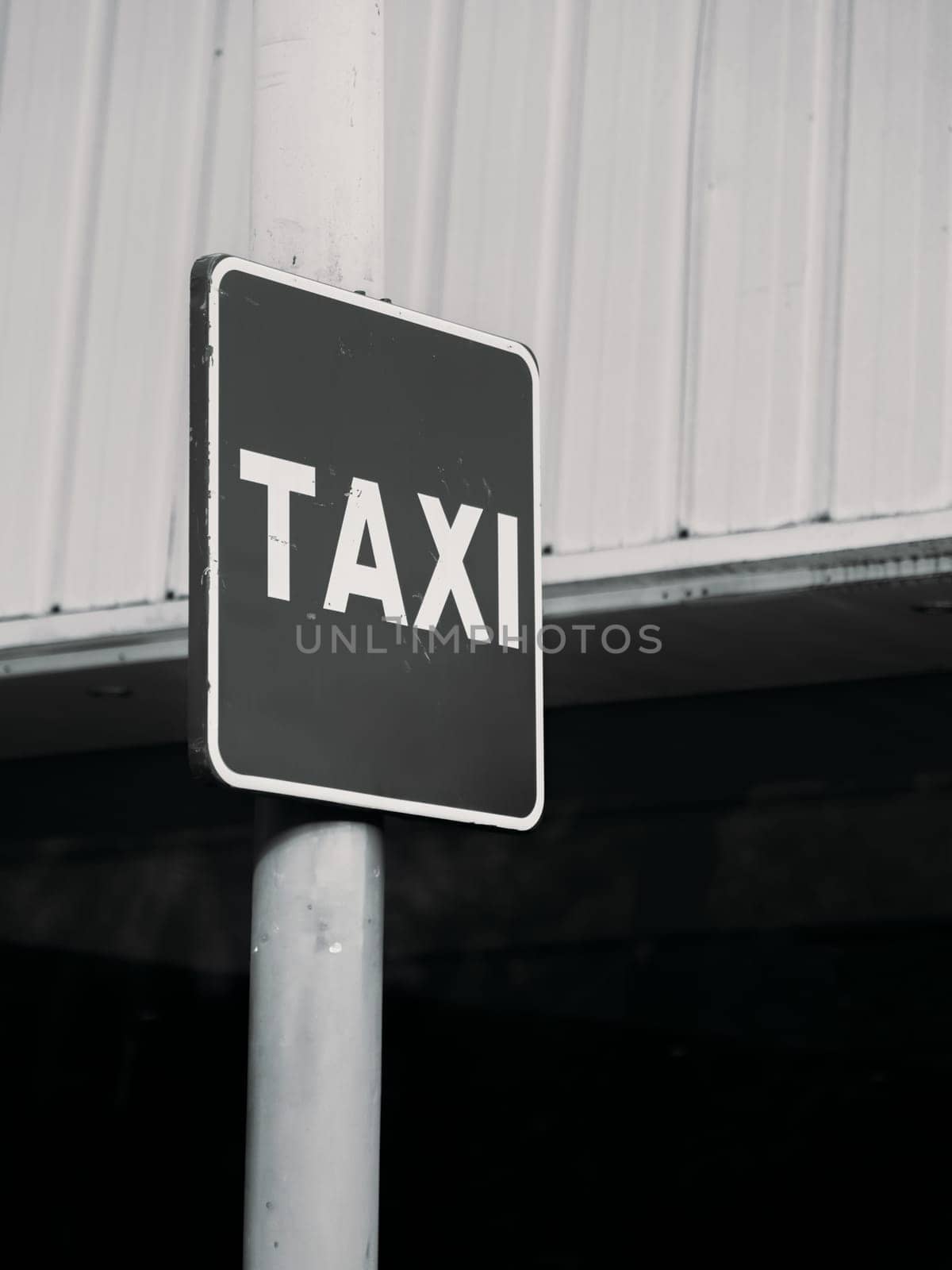 A black and white photo of a taxi sign affixed to a pole with an industrial metal backdrop