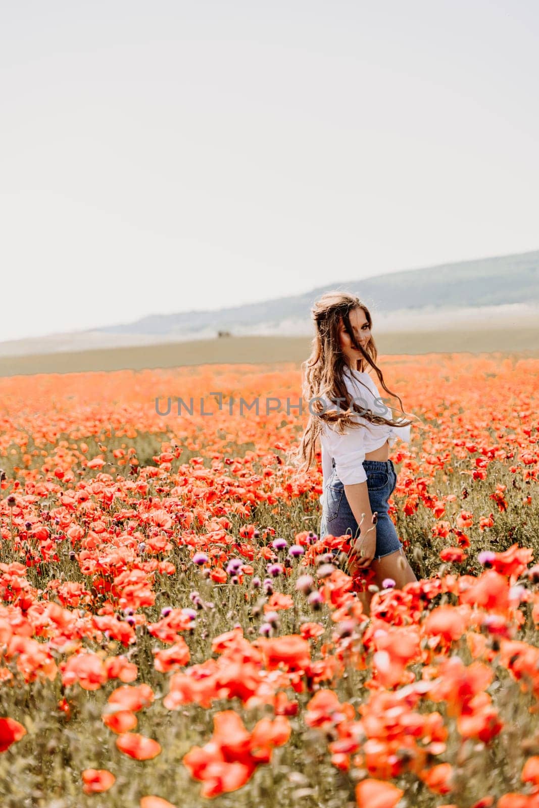 Happy woman in a poppy field in a white shirt and denim skirt with a wreath of poppies on her head posing and enjoying the poppy field