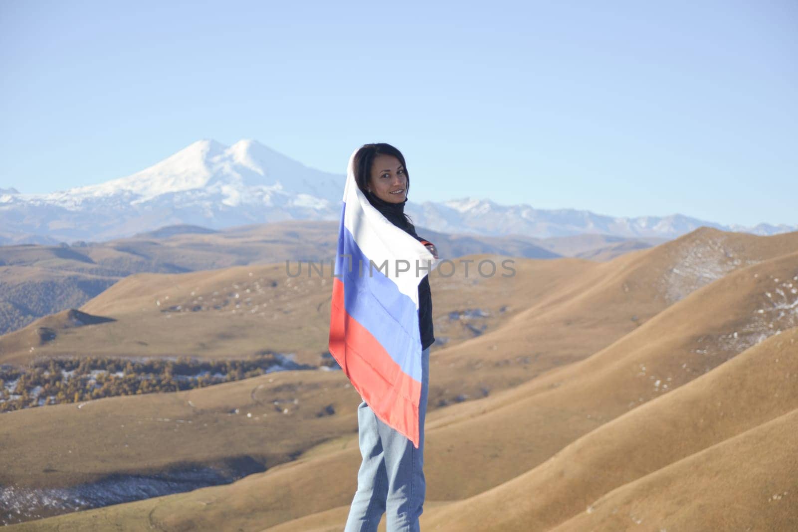 A young brunette woman stands against the backdrop of the snow-capped Mount Elbrus, looking at the camera, a Russian flag covers her shoulders. Tricolor against the backdrop of snow-capped Mount Elbrus by Ekaterina34