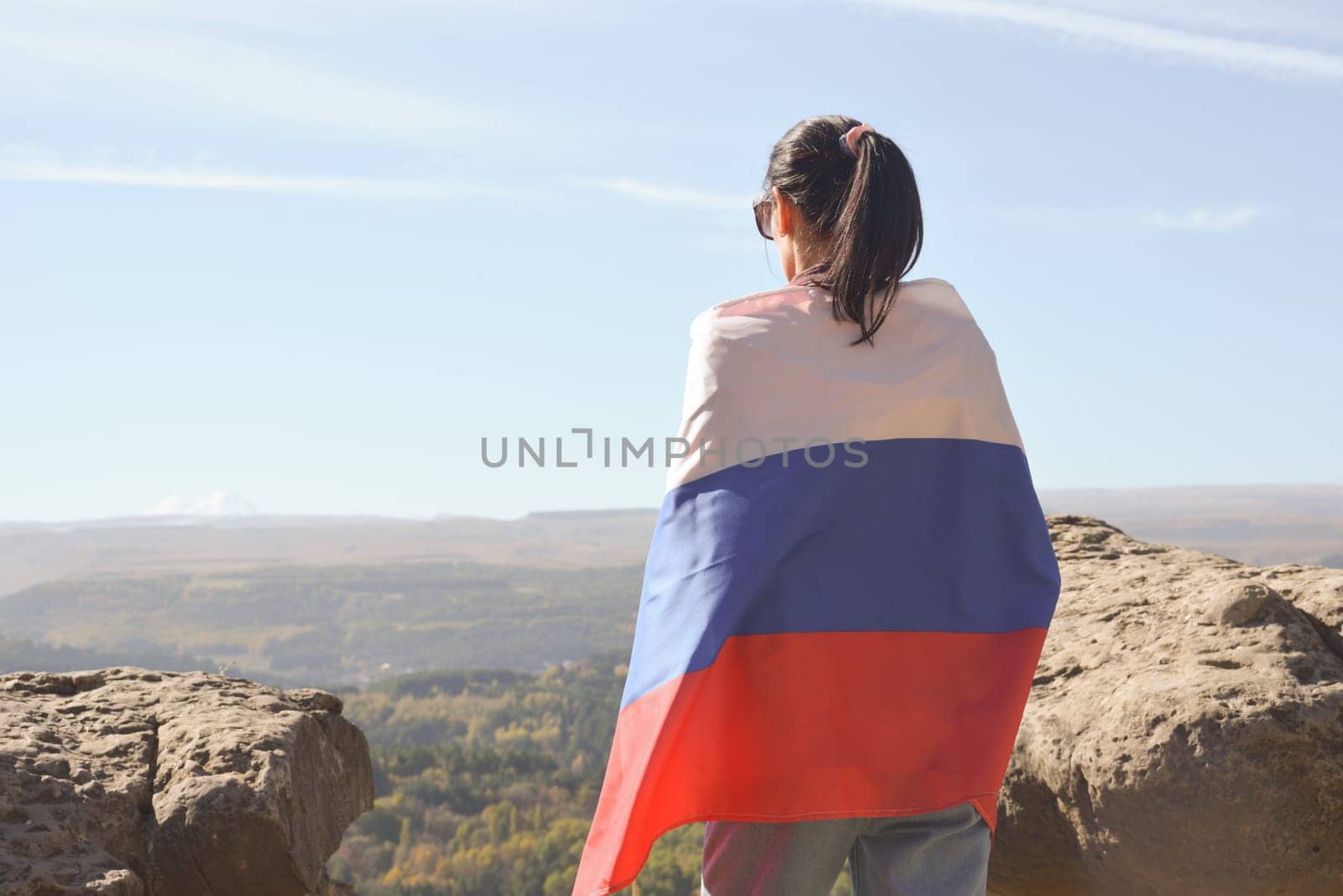 A girl tourist with a Russian flag on her shoulders stands on the top of a mountain and enjoys the beautiful views of the Caucasus Mountains. by Ekaterina34