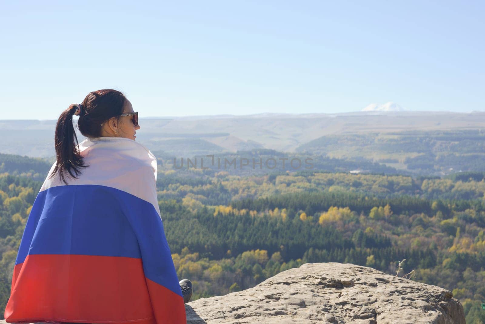 A young brunette woman sits on a stone and looks at the mountains around, her shoulders are covered with a Russian flag