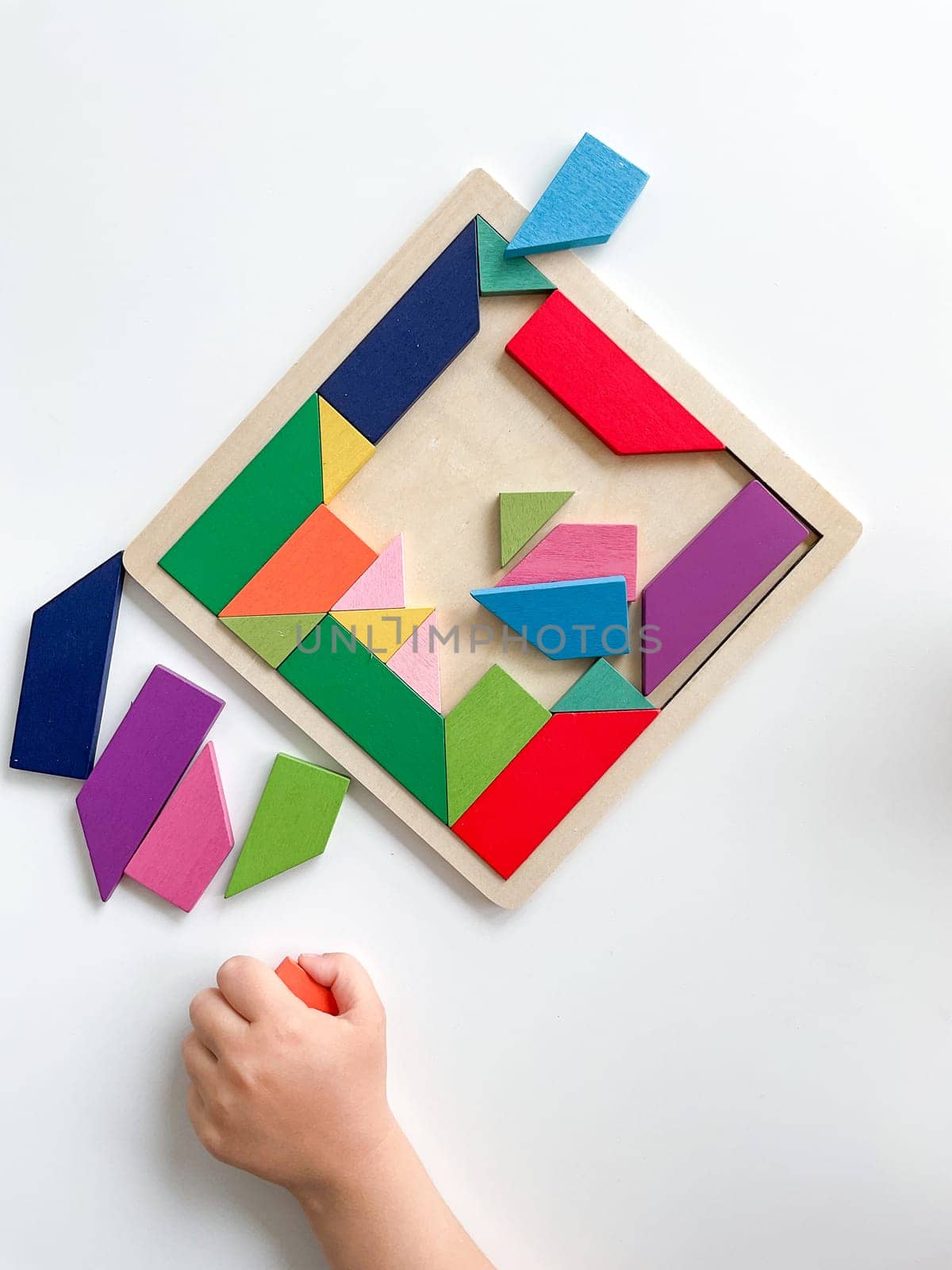 child's hand collects multicolored wooden mosaic on white background. child solves a colorful tangram. square of colorful geometric shapes on white background.