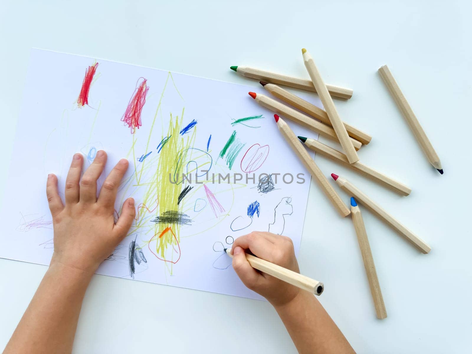 small child draws with colored pencils on paper on white table. by Lunnica