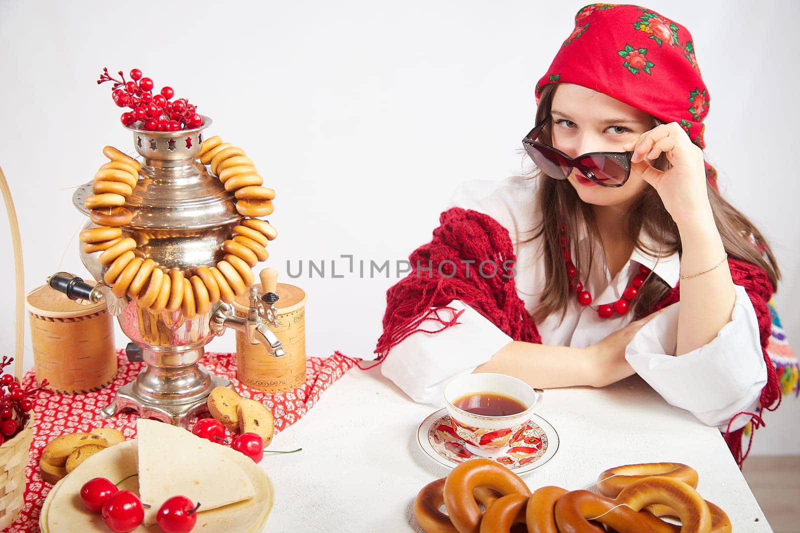 A fashionable modern girl in stylized folk clothes at table with a samovar, bagels and tea for the Orthodox holiday of Maslenitsa and Easter. Funny photo shoot for a young woman