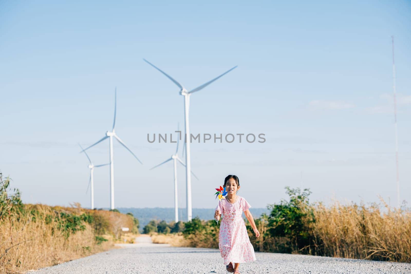 Little girl's playful learning near windmills holding pinwheels with joy. Embracing wind energy education in a cheerful clean electricity generating wind turbine landscape.