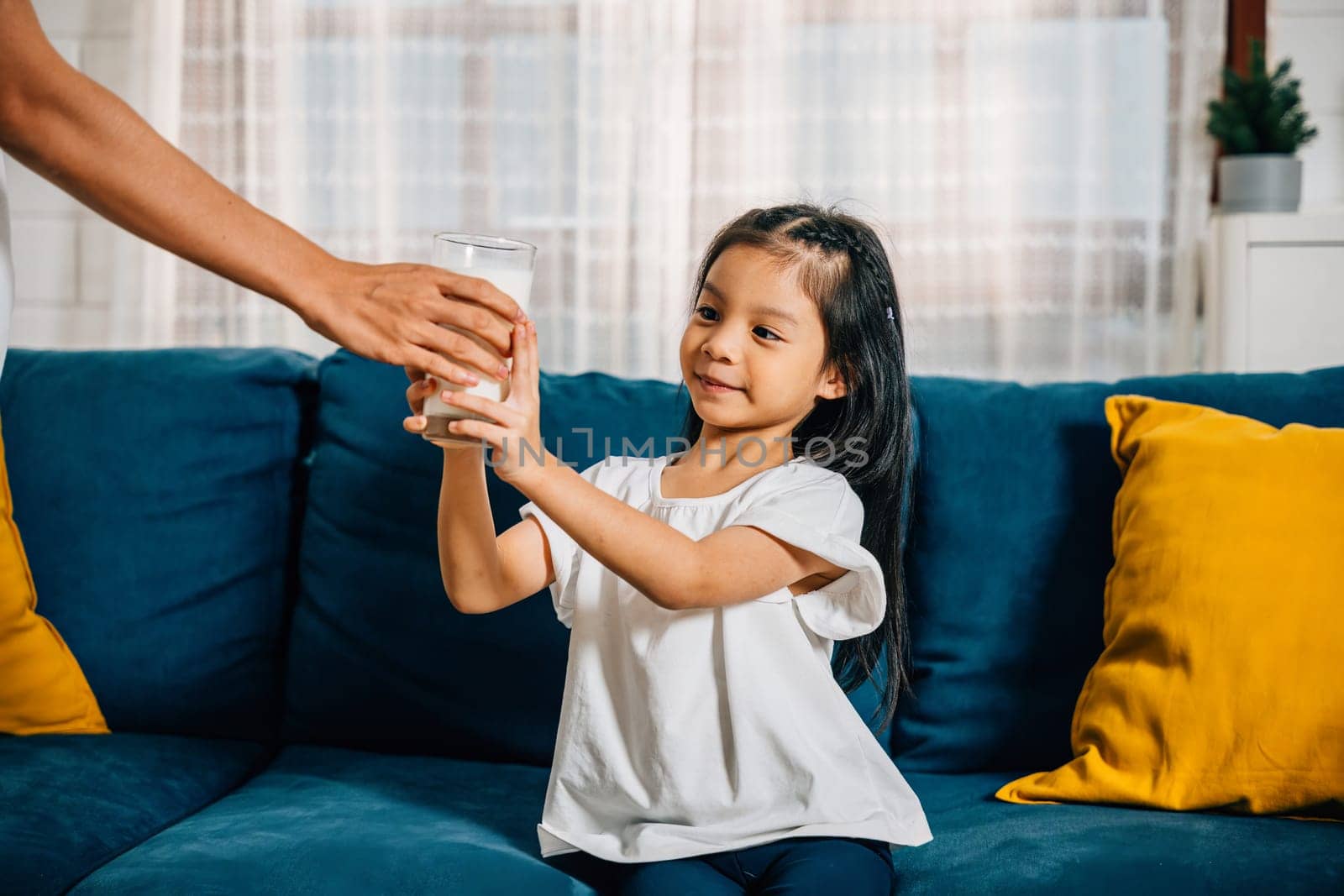 A beautiful family morning as an Asian mother gives her daughter a glass of milk on the sofa creating a picture of innocent joy togetherness and calcium-rich nourishment.