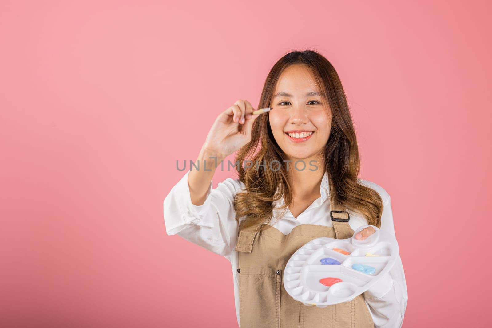 Happy female painting using paintbrush and palette with colors, Asian beautiful young woman artist holding brush and paint palette, studio shot isolated on pink background, Paintings and art equipment