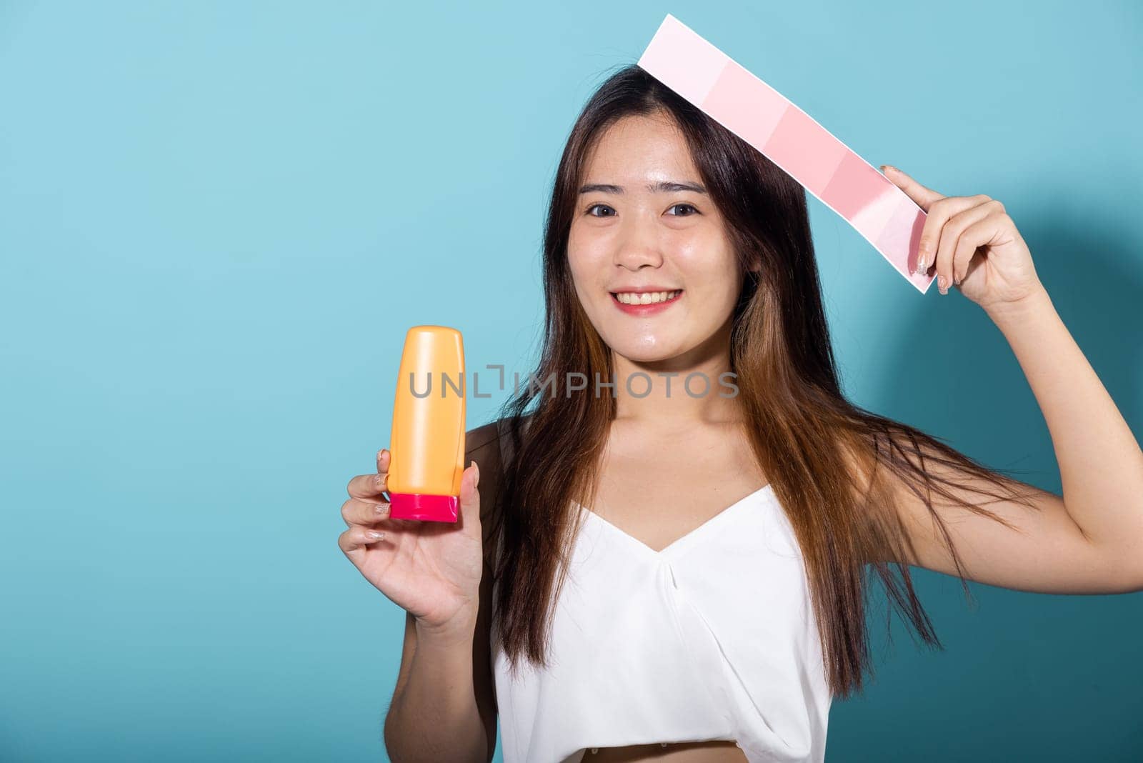 Portrait of happy female hold color card under sun protection and sun cream, Asian beautiful young woman smiling and holding skin color scale paper with suntan lotion isolated on blue background