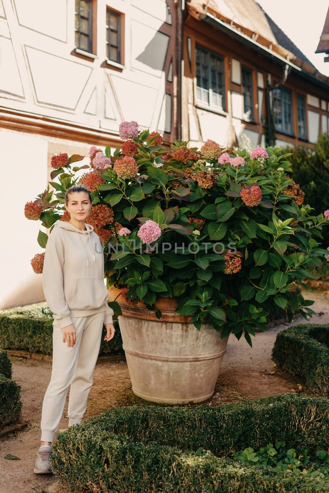 Attractive curly blonde woman walk on the city park street. Girl wear purple hoodie look happy and smiles. Woman make here me gesture standing near pink blooming bush flowers. Happy laughing girl