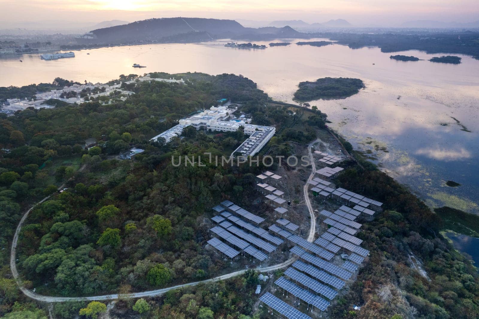 Aerial drone shot with solar panels placed in middle of trees on edge of lake reflecting sunrise colors and aravalli hills showing grene ecological renewable energy production Udaipur by Shalinimathur