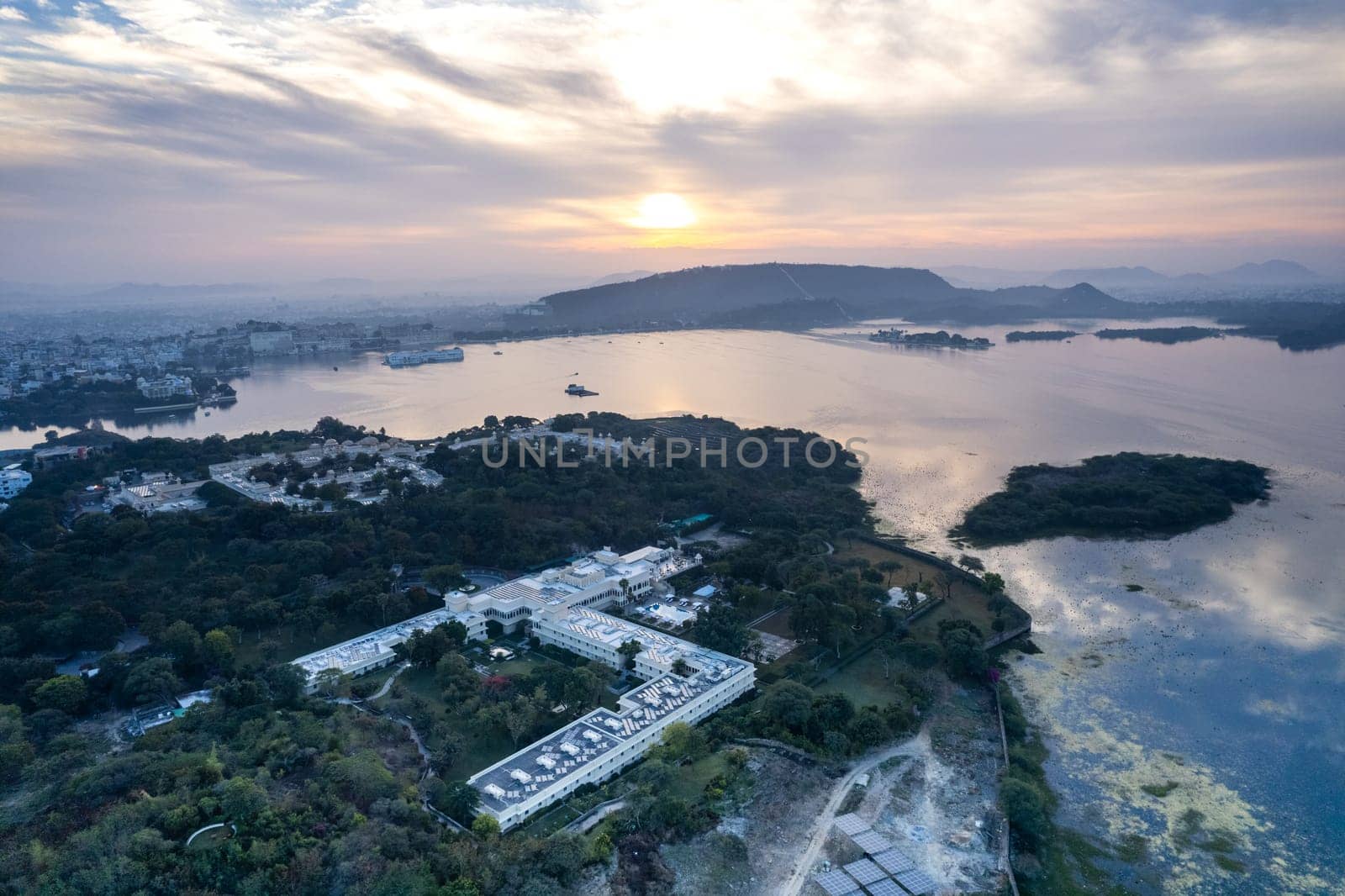 Aerial drone shot showing large resort hotel on tree covered bank facing lake pichola fateh sagar with sun rising over aravalli hills udaipur, Jodhpur by Shalinimathur