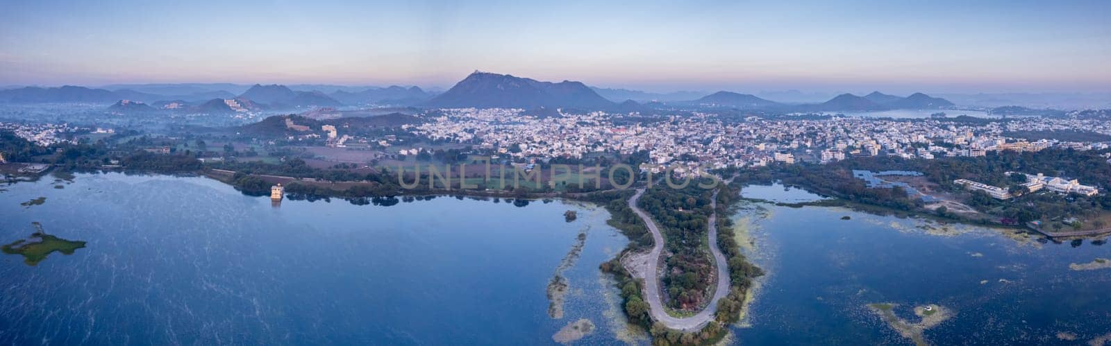 Panoramic aerial drone shot at dawn dusk with road loop extending into fateh sagar lake with aravalli hills in distance hidden in fog showing cityscape of Udaipur Rajasthan India