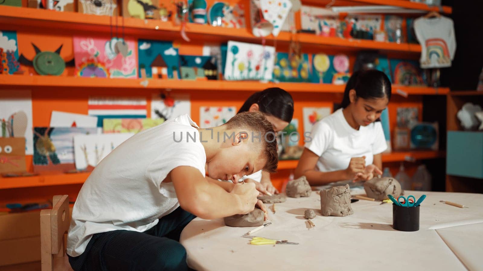 Highschool teenager using carving tool working at clay at pottery workshop. Group of happy diverse children working or modeling cup of clay with dough and equipment scatter around. Edification.