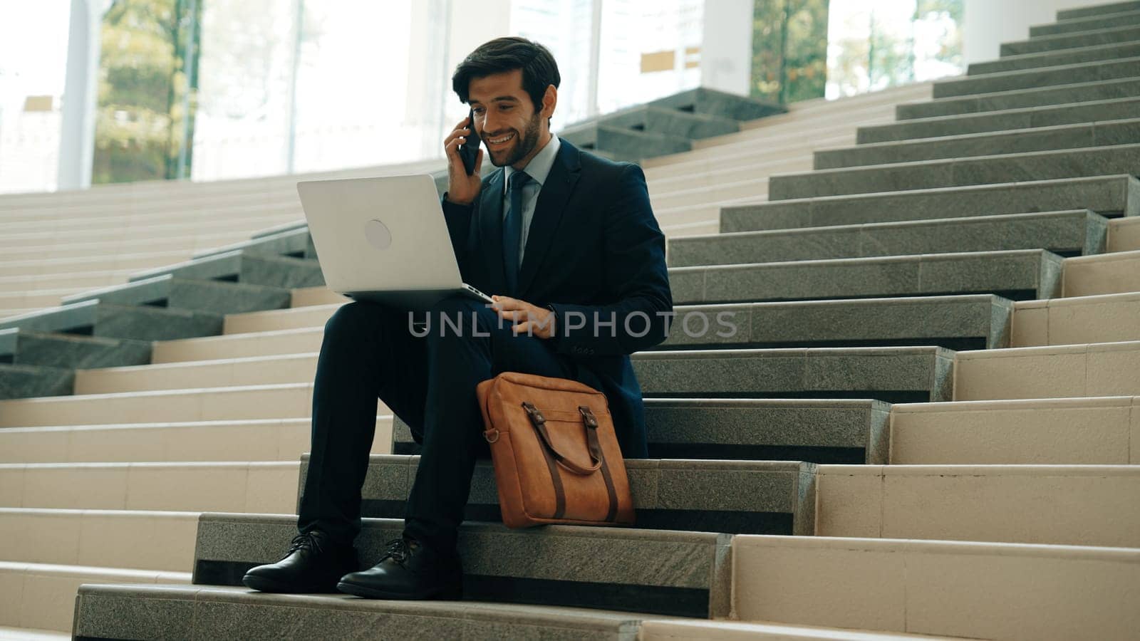 Skilled investor working or planing strategy by using laptop at stair. Professional business man wearing suit while working and typing data analysis by using laptop and talking to phone. Exultant.