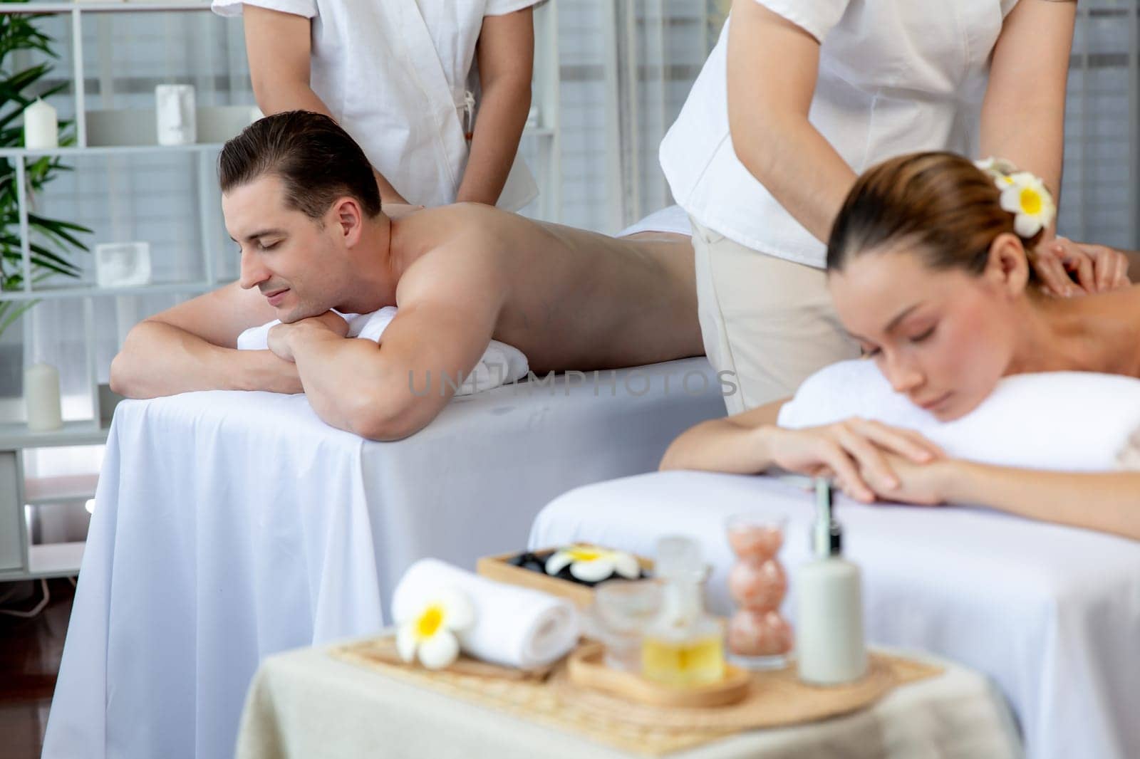 Caucasian couple customer enjoying relaxing anti-stress spa massage and pampering with beauty skin recreation leisure in day light ambient salon spa at luxury resort or hotel. Quiescent