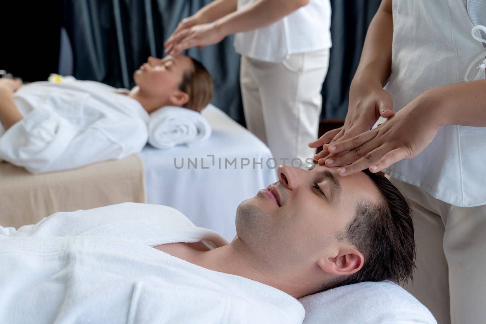 Caucasian couple enjoying relaxing anti-stress head massage and pampering facial beauty skin recreation leisure in dayspa modern light ambient at luxury resort or hotel spa salon. Quiescent
