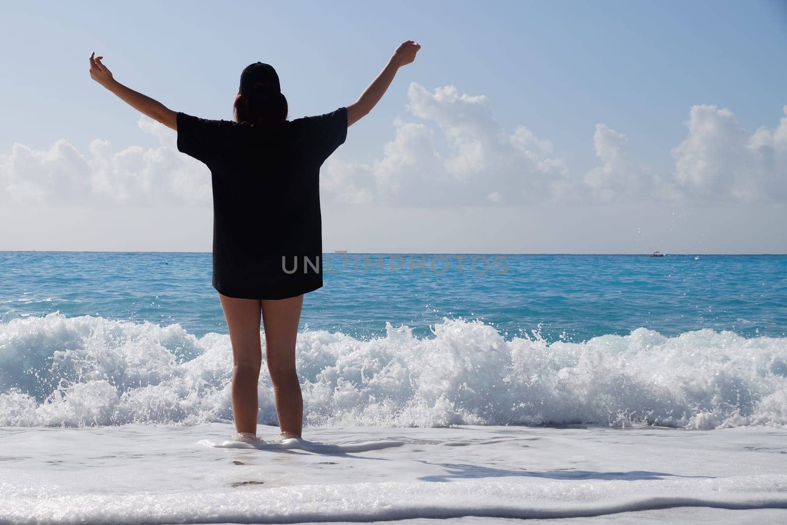 teenage girl in a black T-shirt stands in the sea with her arms outstretched high, rear view.