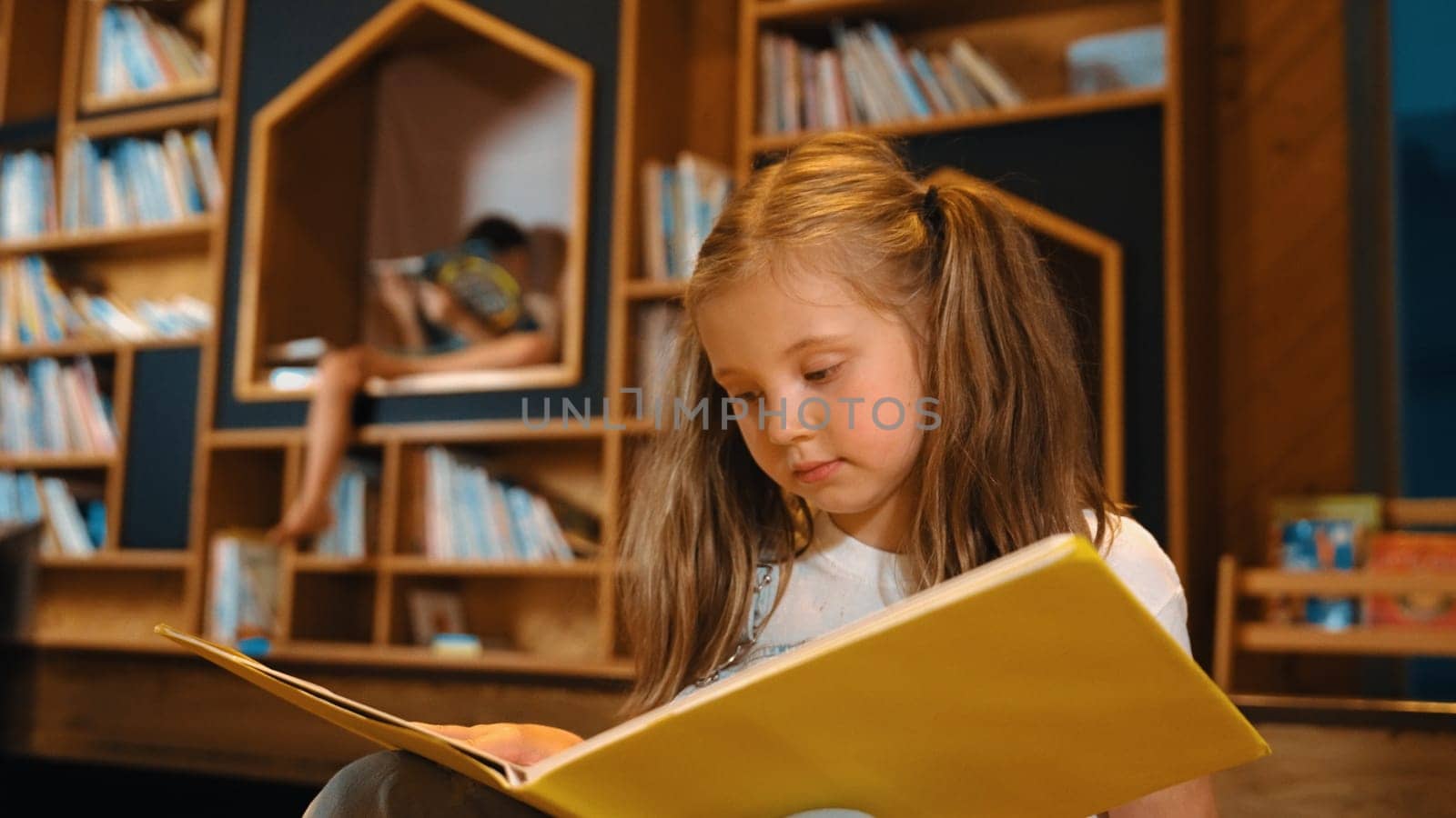 Young smart caucasian girl picking reading a book while sitting at library. Clever child learning, studying, open a books at library. Attractive kid turning page with blurring background. Erudition.