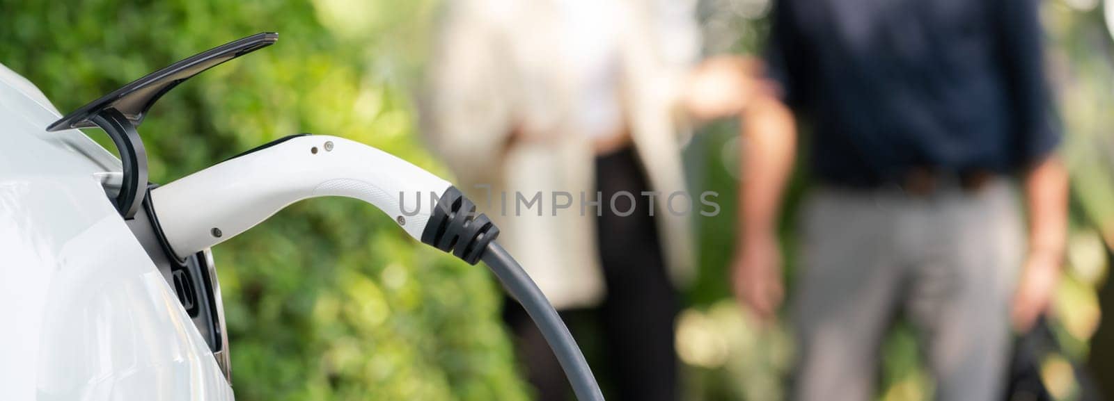 Young couple recharge electric car battery from charging station in green city park in springtime. Rechargeable EV car for sustainable environmental friendly urban travel lifestyle. Panorama Expedient