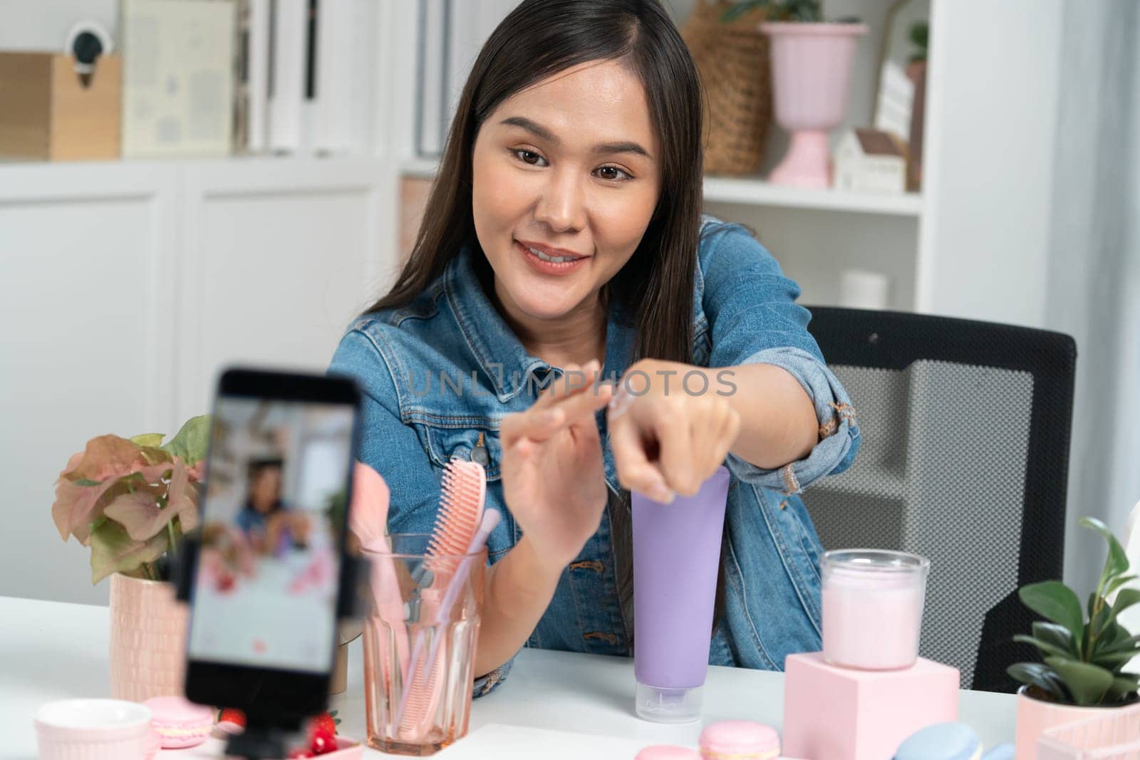 Young beautiful Asian girl reviewing moisturizing body lotion cream product to make glow skin testing on arm on social media online live recording smartphone with bestseller at modern room. Stratagem.