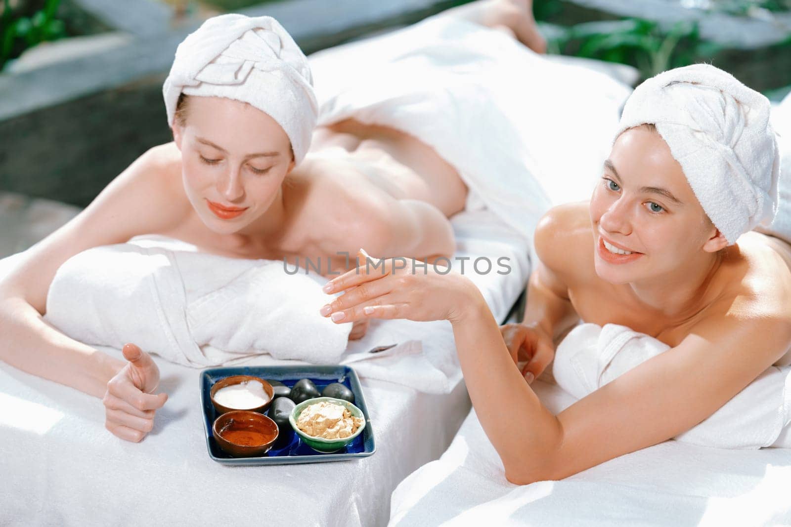 Couple of beautiful young girls lie on spa bed during interested in homemade beauty facial mask. Attractive woman touching herbal facial mask. Surrounded with nature environment. Tranquility.