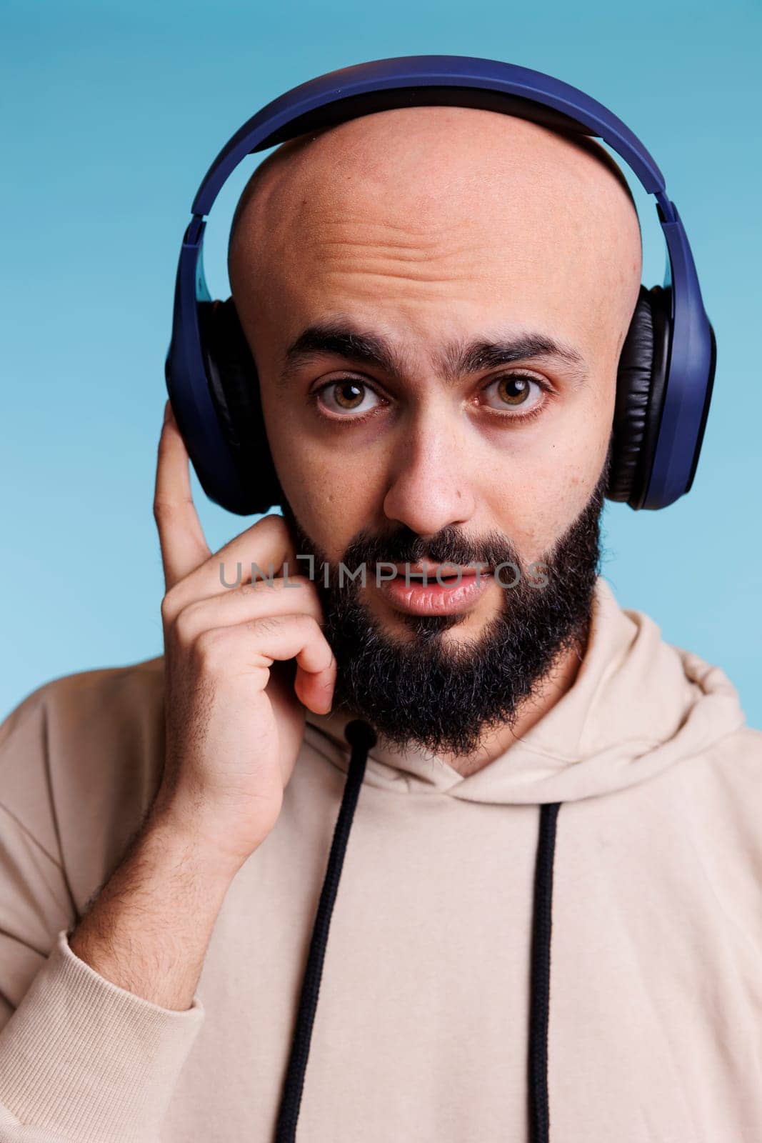 Young arab man listening to music in wireless headphones portrait. Bald bearded person enjoying tunes playlist in earphones while looking at camera with neutral expression