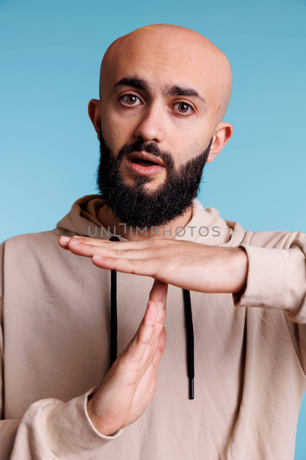 Arabian person taking time out with stop signal while showing confident facial expression portrait. Young bald bearded man making interruption gesture with hands while looking at camera