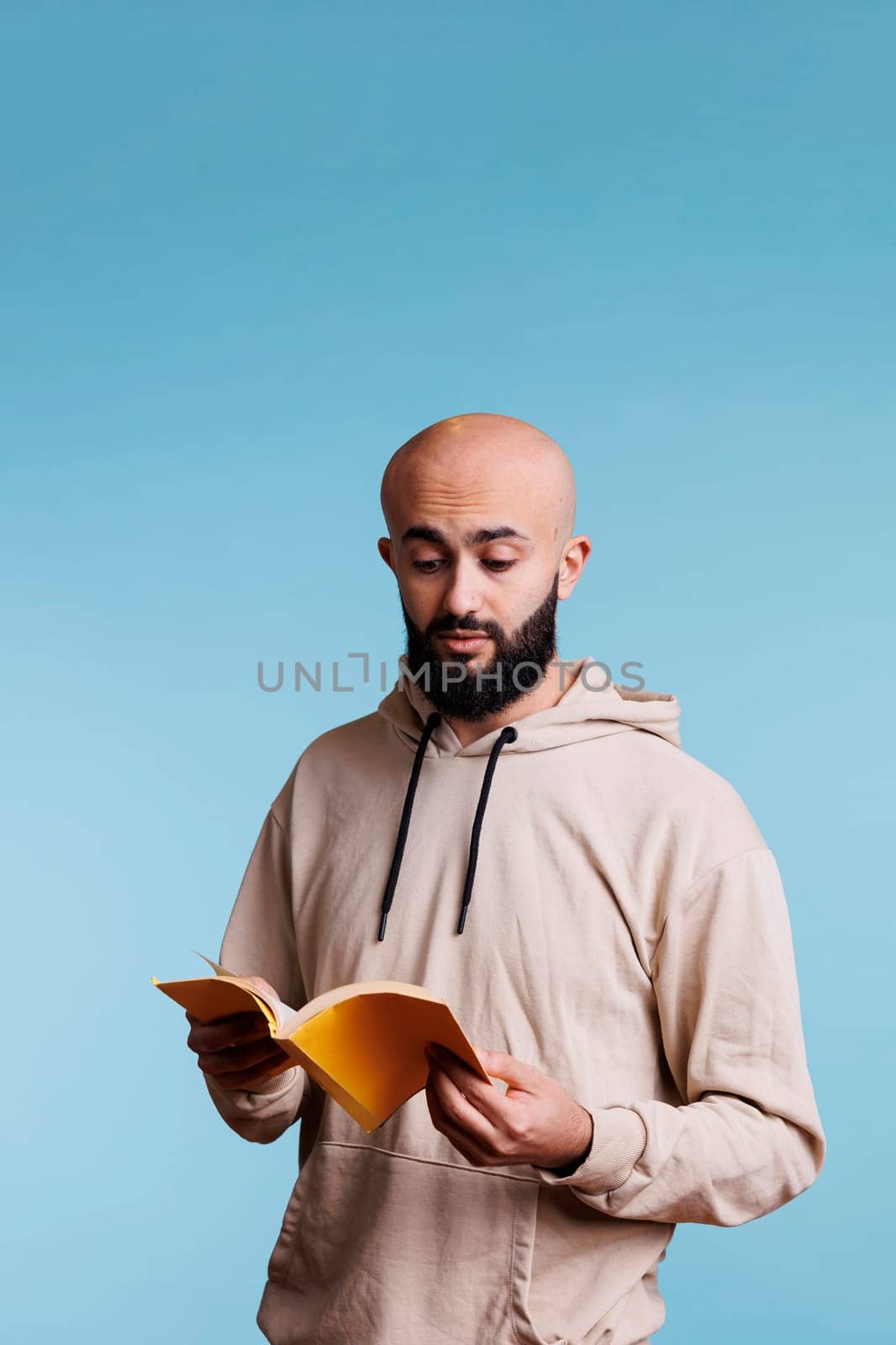 Arab man reading book paperback edition with yellow cover. Young concentrated bald bearded person in hoodie holding open notepad while standing with surprised facial expression