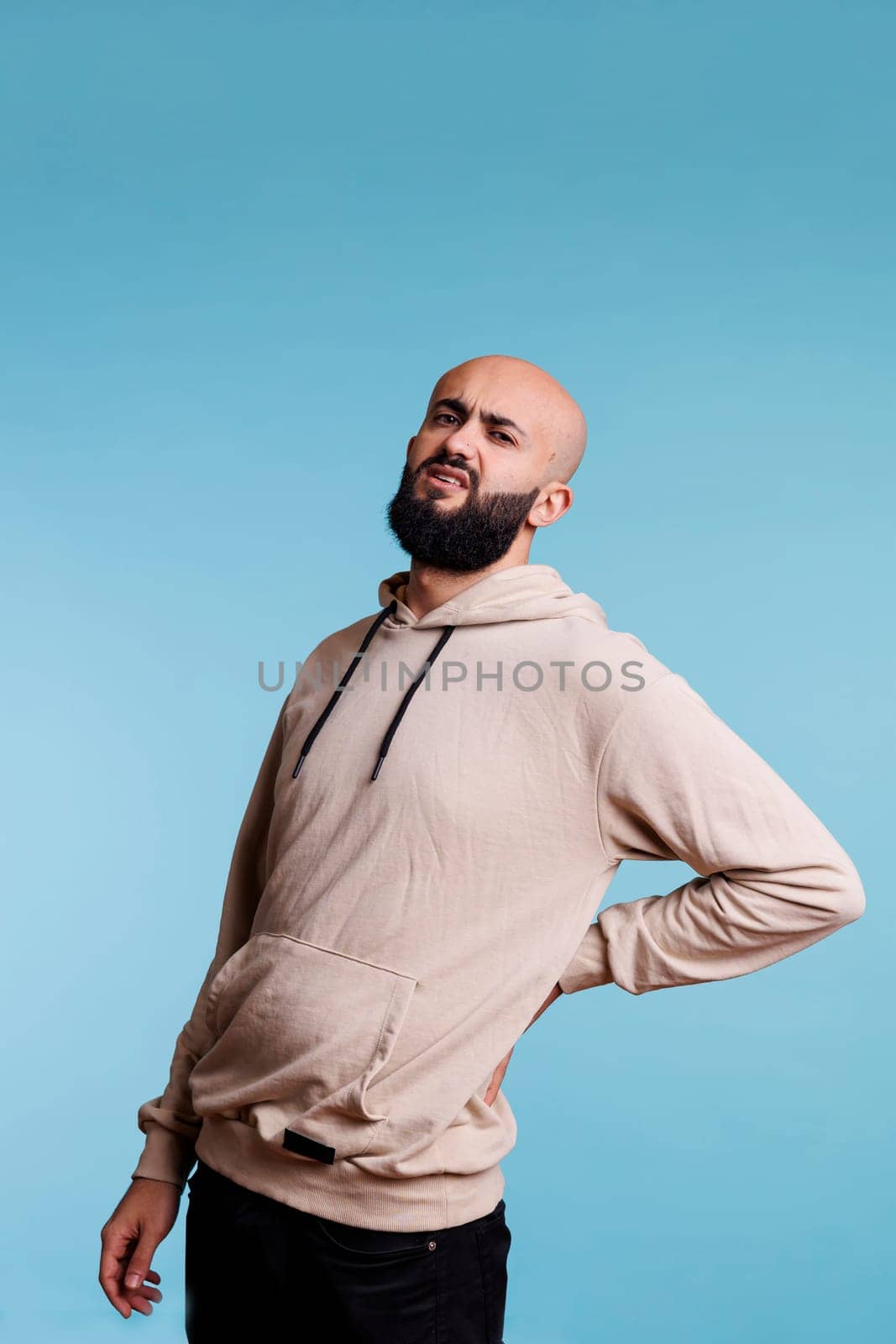 Arab man grimacing in pain while holding lower back and looking at camera. Young person wearing casual clothes suffering from chronic backache and muscles cramps portrait
