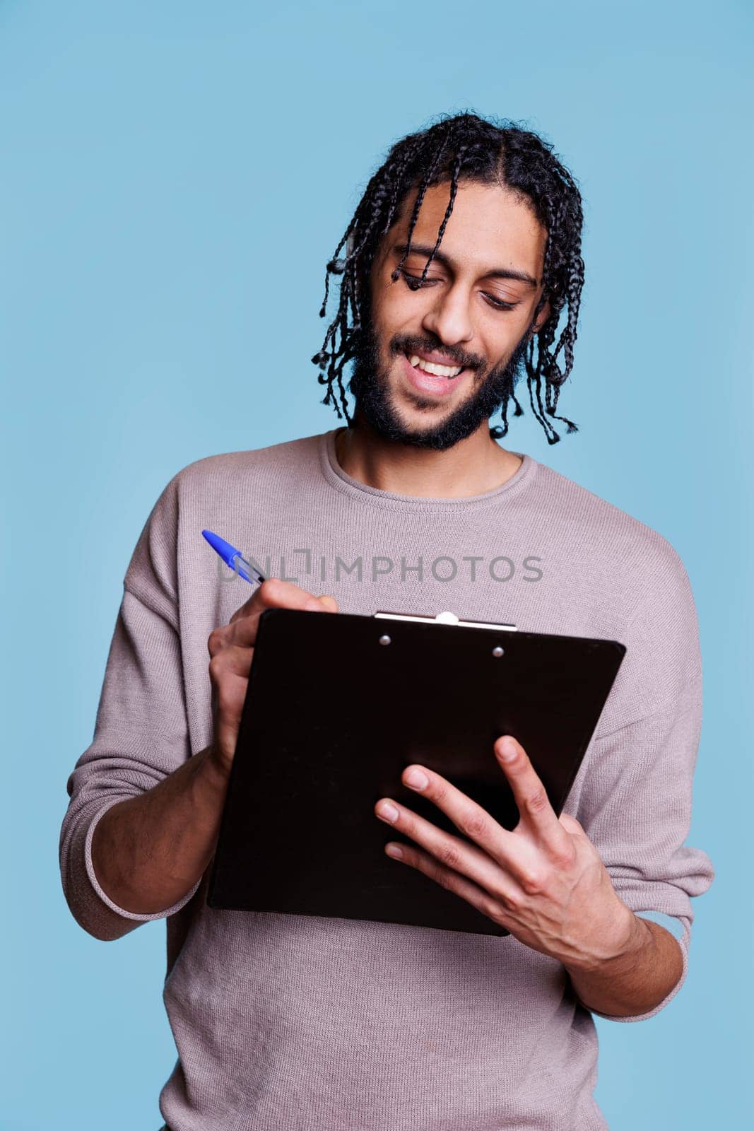 Smiling arab man holding clipboard and writing plan checklist with cheerful facial expression. Carefree young person wearing casual clothes taking notes and filling application