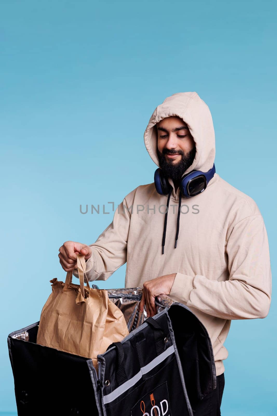 Arab food service deliveryman in hood taking package with lunch from backpack. Young bearded man wearing casual beige hoodie holding paper bag with takeaway dish and thermal bag