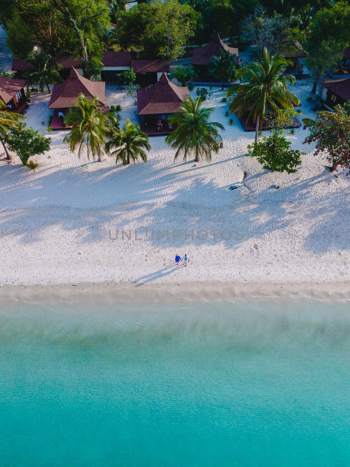 Drone view at a couple walking on the white sandy tropical beach of Koh Muk with palm trees soft white sand, and a turqouse colored ocean in the evening in Koh Mook Trang Thailand