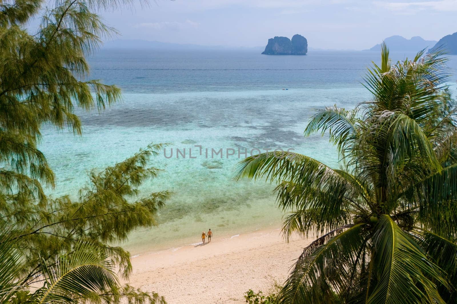 view behind palm trees from a drone at a couple on the beach of Koh Ngai Island Thailand, with palm trees and soft white sand, and a turqouse colored ocean in Koh Ngai Trang Thailand