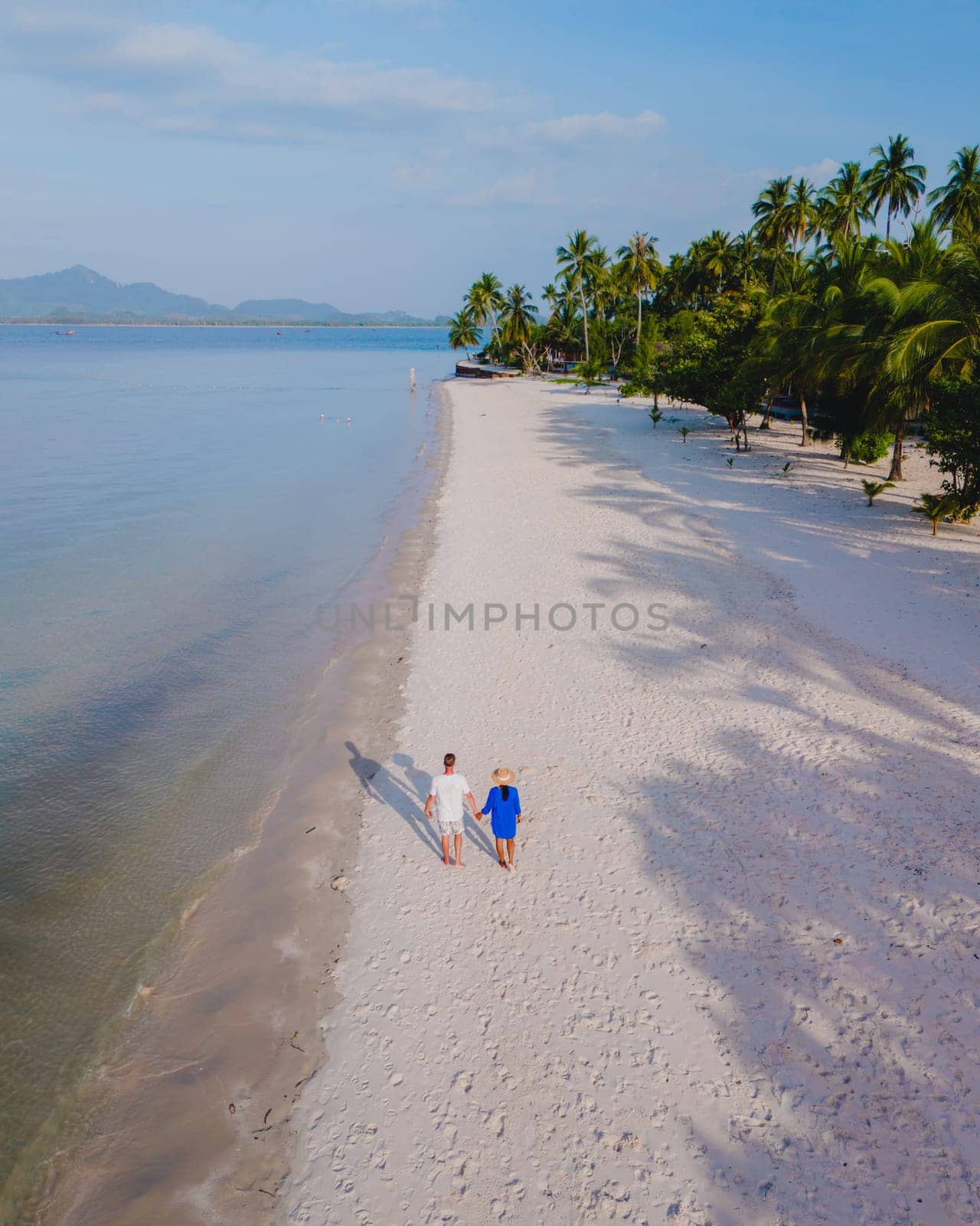 Drone view at a couple walking on the white sandy tropical beach of Koh Muk with palm trees soft white sand, and a turqouse colored ocean in Koh Mook Trang Thailand on a sunny afternoon