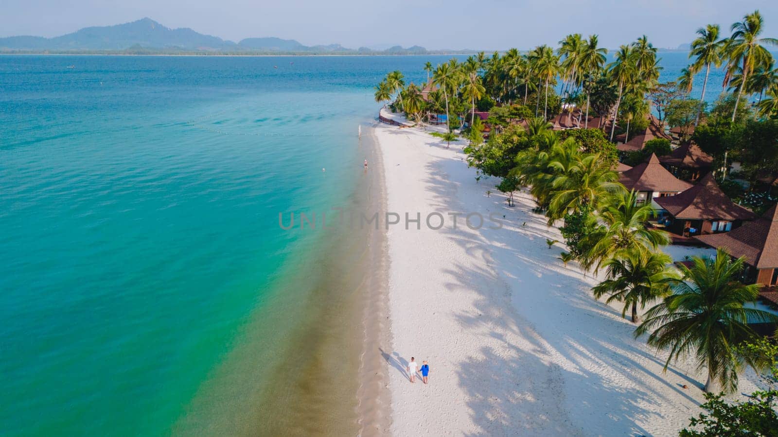 Drone view at a couple walking on the white sandy tropical beach of Koh Muk with palm trees soft white sand, and a turqouse colored ocean in Koh Mook Trang Thailand on a sunny afternoon