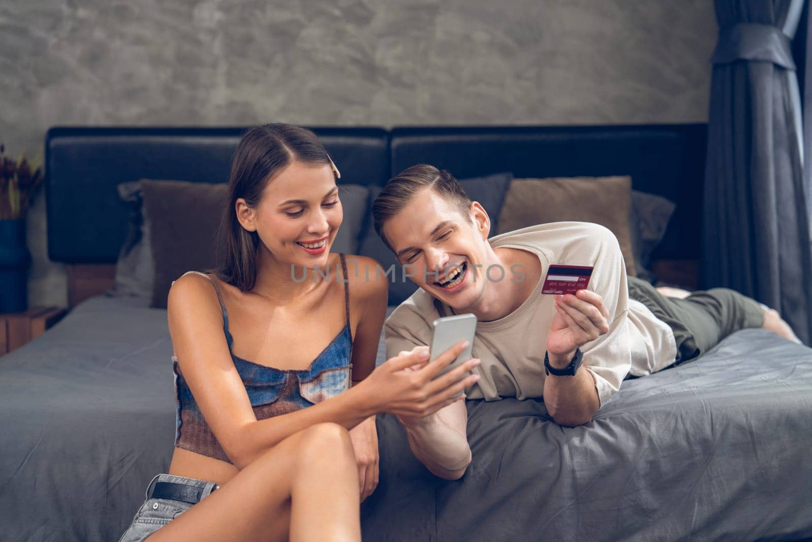 Young couple sit in the home bedroom using online payment app and digital wallet on smartphone to pay with credit card. E commerce shopping and modern purchasing via mobile internet. Unveiling