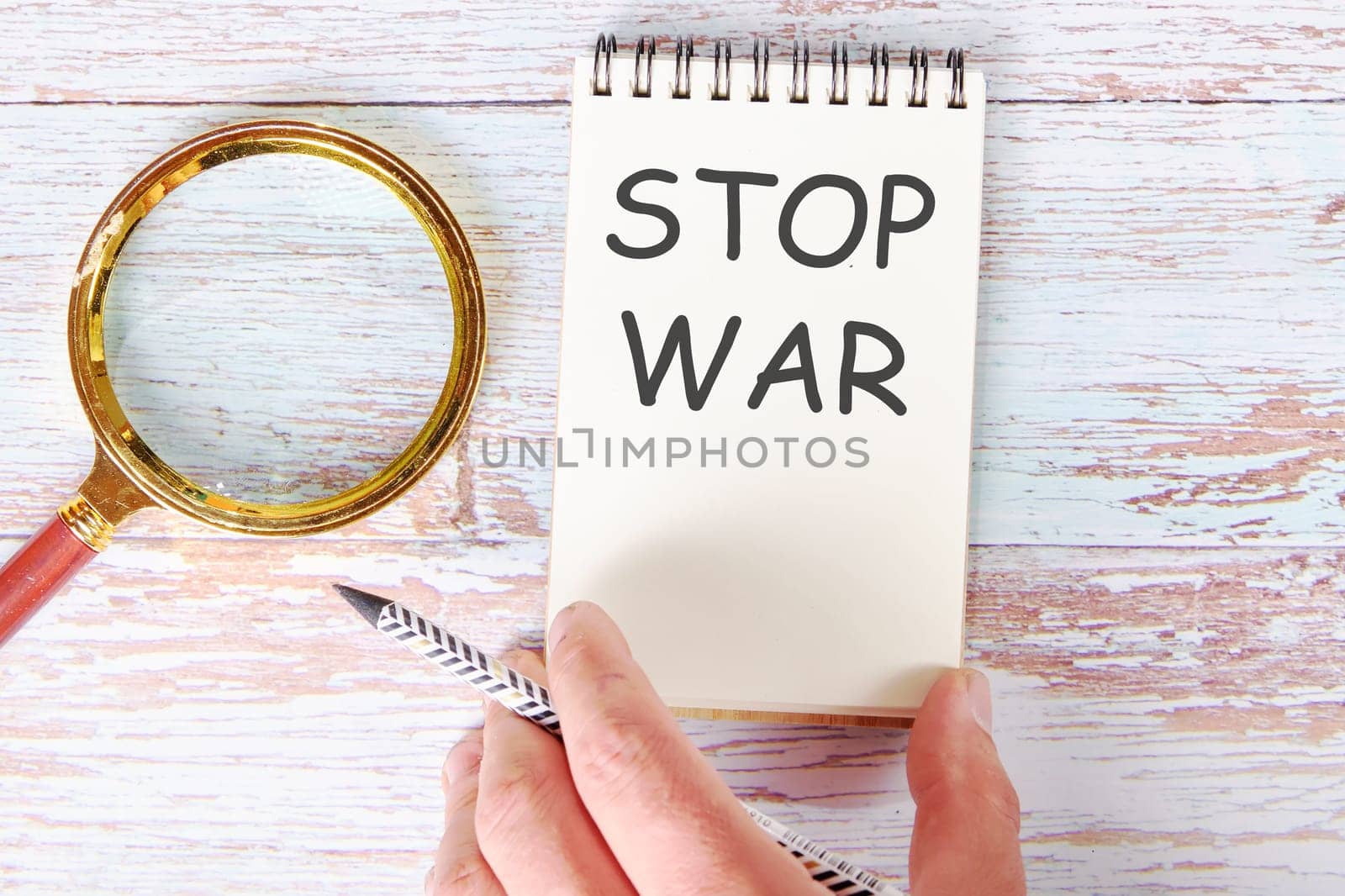 STOP WAR written on a blank sheet of a notebook which is touched by a man's hand with a pencil and a magnifying glass is lying next to it