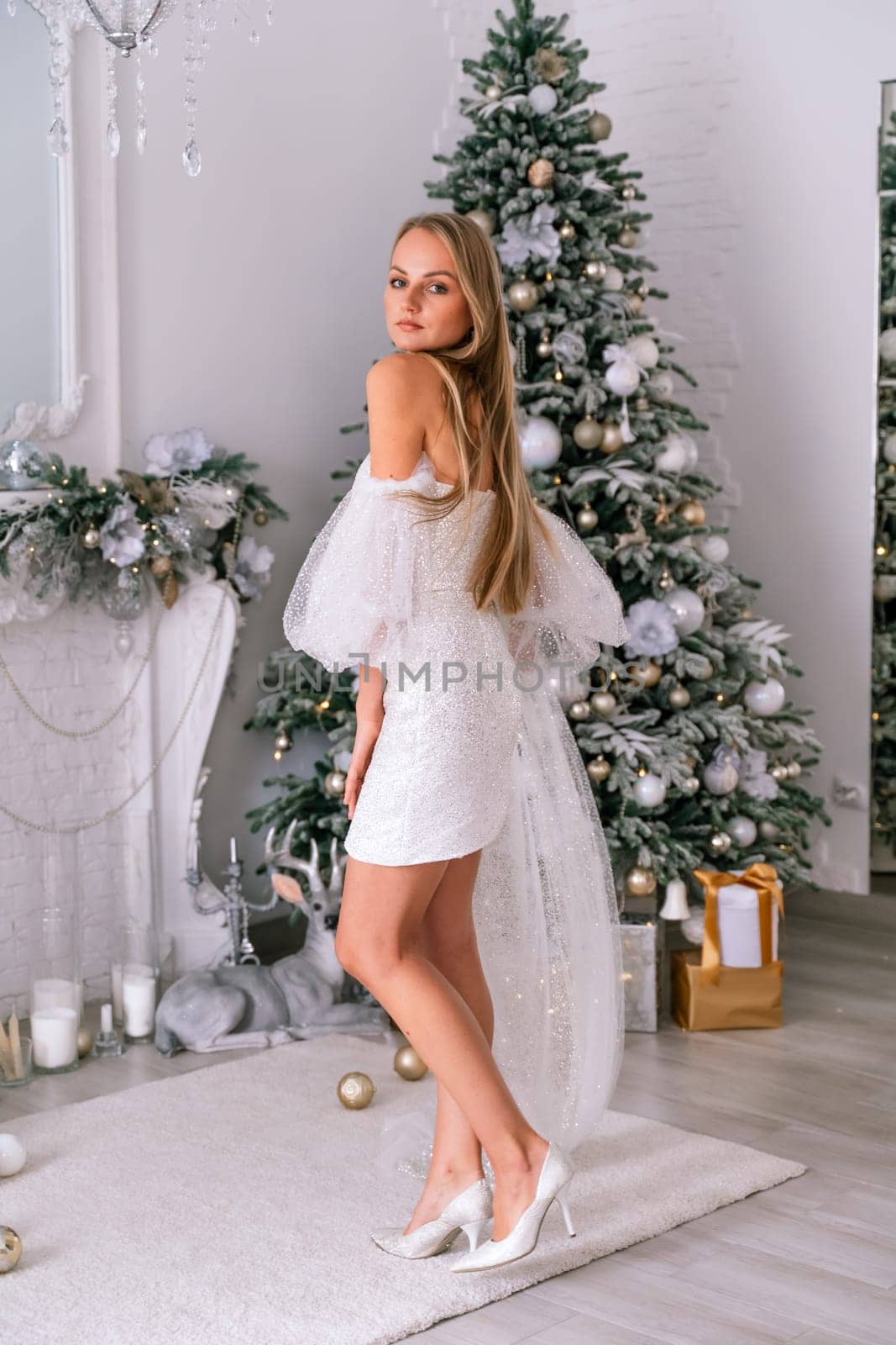 The blonde in the Christmas room. A beautiful blonde woman in a shiny light short dress with a train stands in a beautiful bright room decorated with a festive interior with a Christmas tree