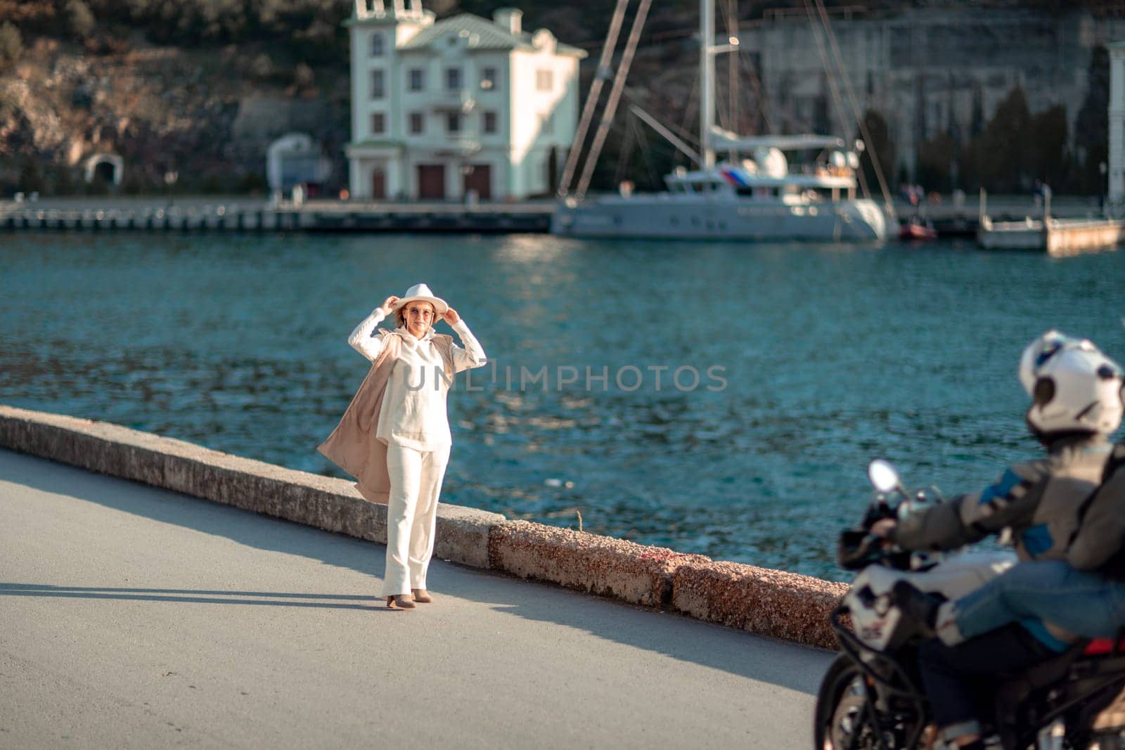 Happy blonde woman in a white suit and hat posing at the camera against the backdrop of the sea.