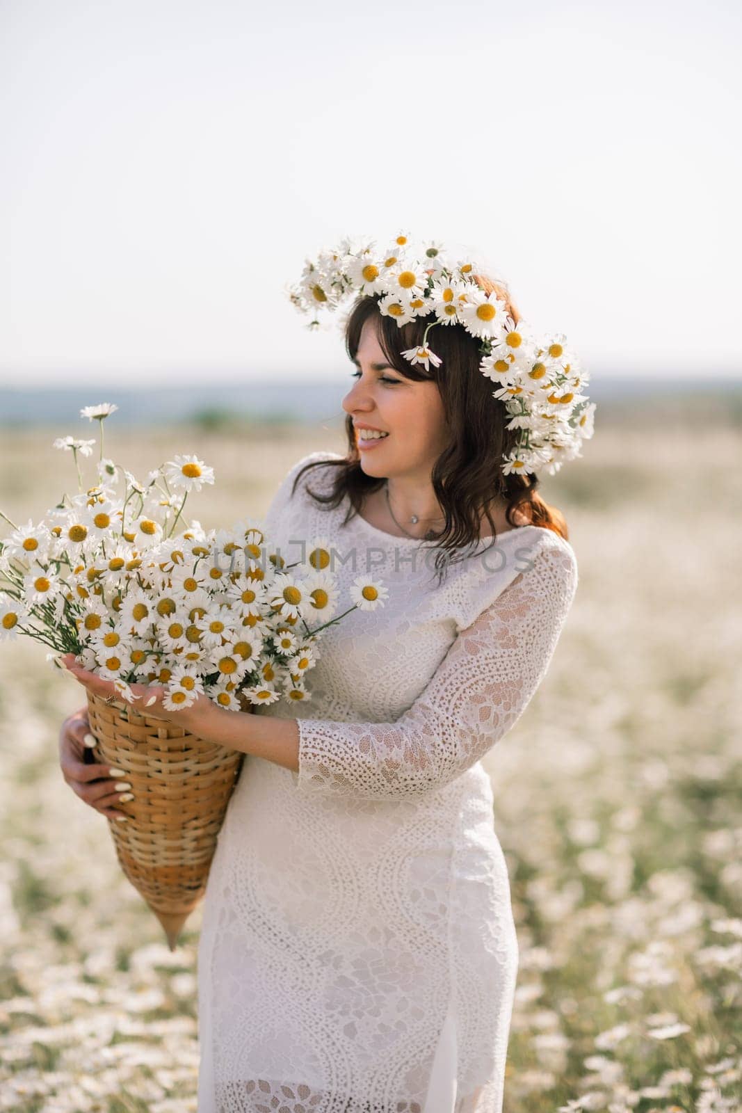 Happy woman in a field of daisies with a wreath of wildflowers on her head. woman in a white dress in a field of white flowers. Charming woman with a bouquet of daisies, tender summer photo.