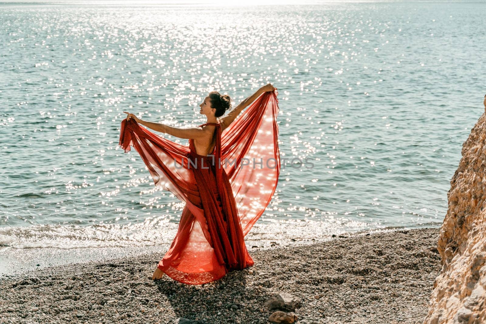 Woman red dress sea. Female dancer in a long red dress posing on a beach with rocks on sunny day. Girl on the nature on blue sky background