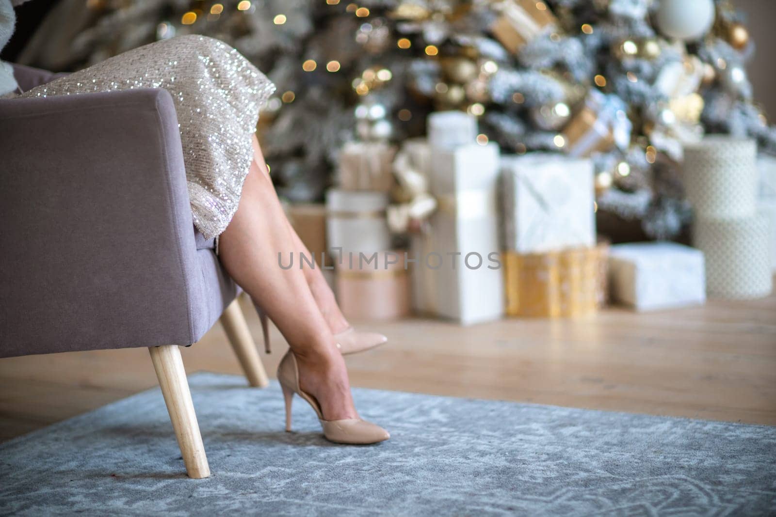 Women's legs. Cropped close-up photo of healthy beautiful elegant female legs in beige high-heeled shoes, woman sitting in a chair on the background of a Christmas tree with gifts.