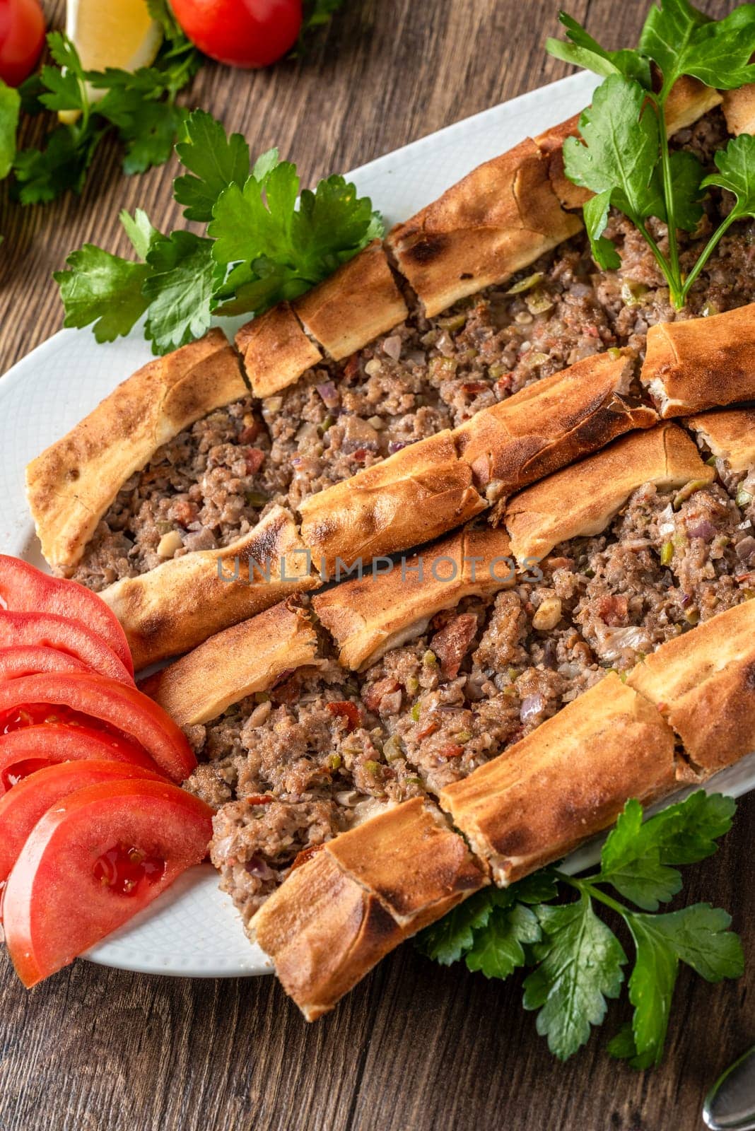 Traditional Turkish dish minced meat pita decorated with tomatoes and greens on wooden table by Sonat