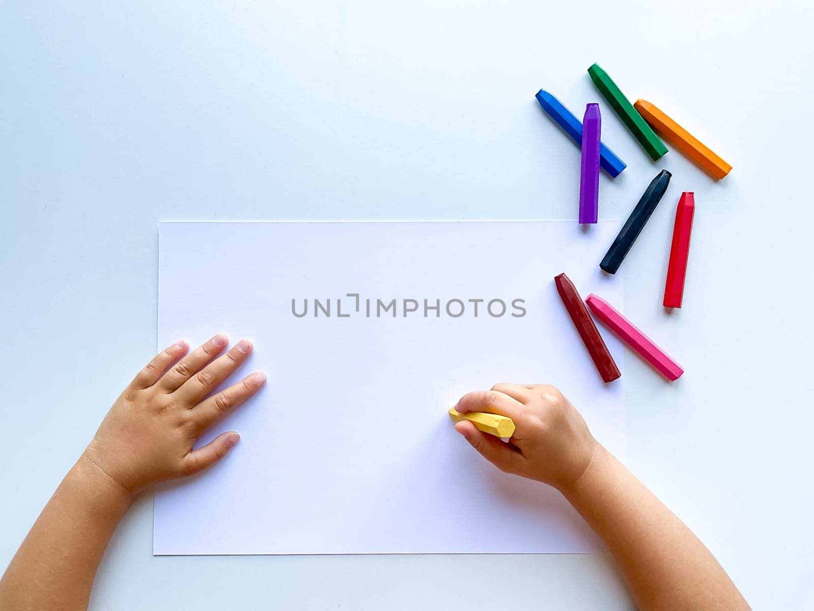 Childrens hands draw with colored wax crayons on a white sheet of paper. Top view of a blank sheet. by Lunnica