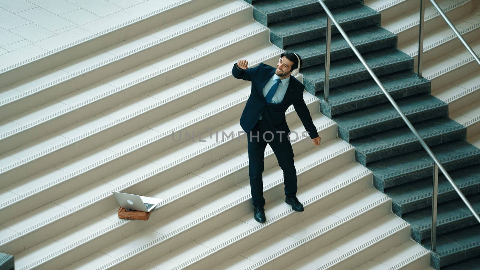 Professional business man standing at stair while moving to music from headphone. Skilled project manager dancing while listening relax sound with laptop placed on stairs. Modern dance. Exultant.