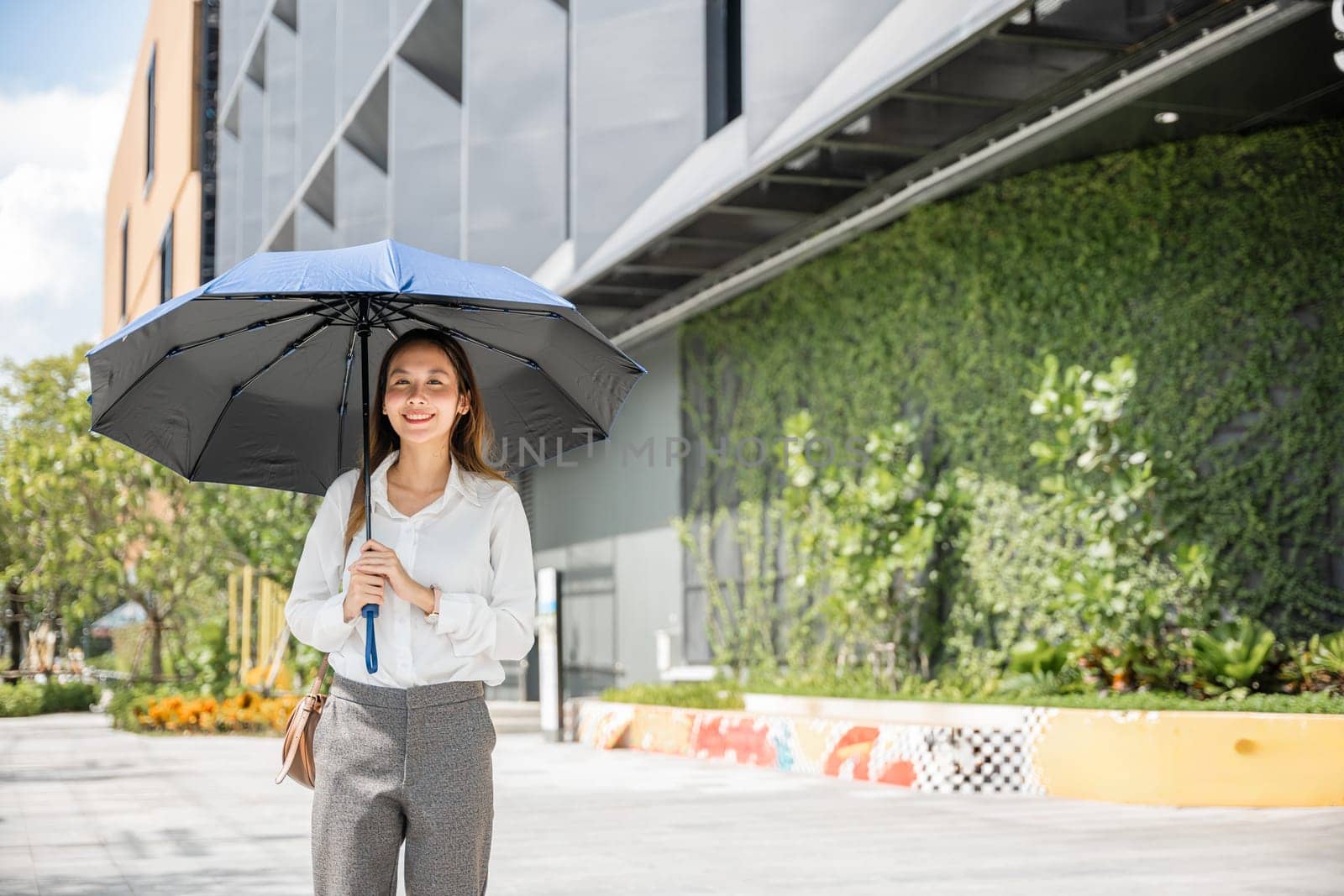 Walking to the office on a scorching day, a young businesswoman holds an umbrella to shield herself from the hot sun. Her determination and sweat highlight her commitment to success. by Sorapop