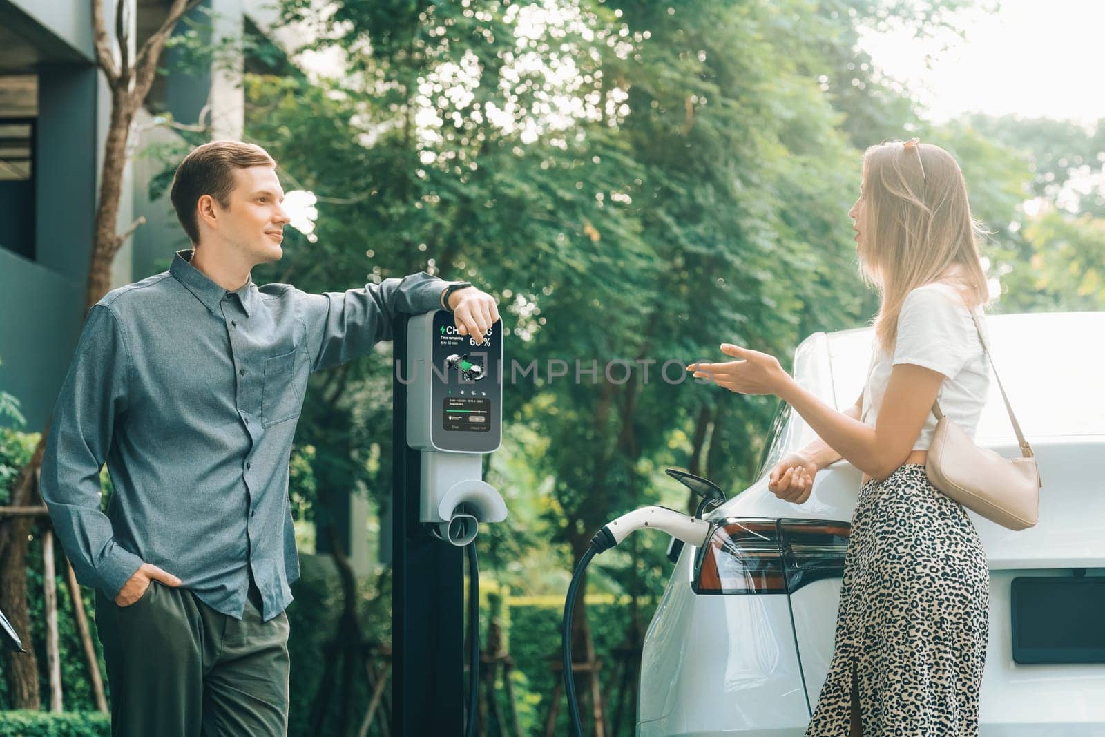 Young couple travel with EV electric car charging in green sustainable city outdoor garden in summer shows urban sustainability lifestyle by green clean rechargeable energy of electric vehicle innards