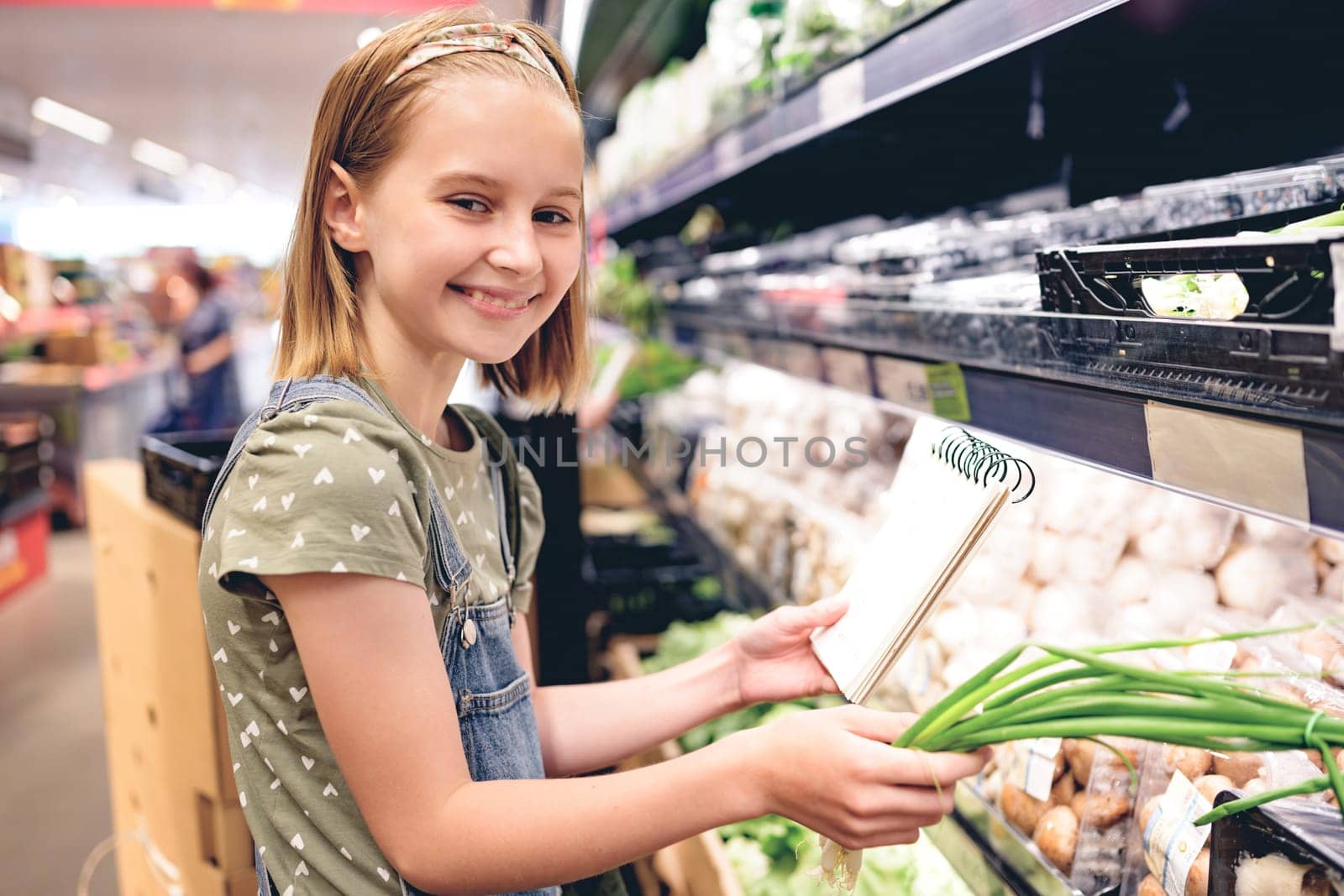 Pretty girl child buying green onion with shopping list in supermarket. Beautiful female preteen kid choosing vegetables in grocery store